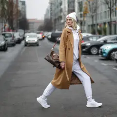 Kobieta w beżowym płaszczu i białych dresach oversize przechodzi przez ulicę, niosąc torebkę.