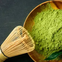 Ein hölzerner Löffel mit feinem, leuchtend grünem Matcha-Pulver und ein Bambusbesen daneben. Der Geschmack ist intensiv, leicht bitter.