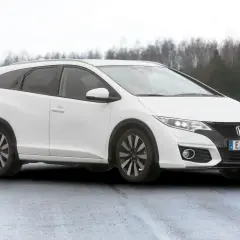 Vit Honda Civic Tourer parkerad på en fuktig väg med träd i bakgrunden.