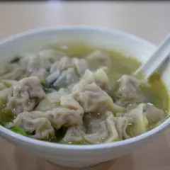 A miseczka pełna aromatycznej zupy wonton, z widocznymi pierożkami i zielonymi warzywami. Łyżka zanurzona w bulionie.