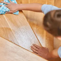 Une main nettoie un escalier en bois verni encrassé avec un chiffon bleu à carreaux.