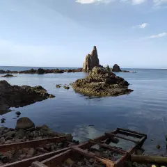 Rocas emergiendo del mar azul, como el arrecife de las sirenas. El agua cristalina revela el lecho rocoso y restos oxidados en la orilla.