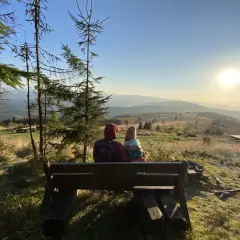 Para siedzi na ławce, podziwiając widok na góry. Słońce zachodzi nad horyzontem, malując niebo na złote i pomarańczowe barwy. To piękny widok z mogielicy z Jurkowa.