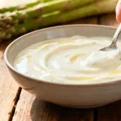 Un bol de sauce hollandaise légère, onctueuse et dorée, avec des asperges vertes floues en arrière-plan sur une table en bois.
