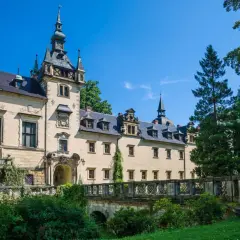 Elegancki hotel zamek z wieżą i mostkiem, otoczony zielenią, pod błękitnym niebem.