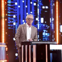 Mężczyzna w garniturze stoi przy pulpicie na tle sceny z niebieskimi światłami.
