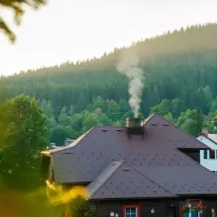 Dym z komina na dachu domu, w tle las i góry. To dobry moment na przegląd kominiarski.