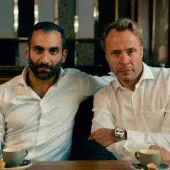Två män i vita skjortor sitter vid ett bord med kaffe. En arm vilar på den andres axel. Scen från hundarna serie.