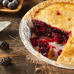 Una fetta di torta di more appena sfornata, con il ripieno succoso che cola. Due more fresche adagiate sul tavolo di legno.