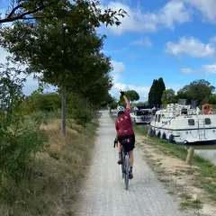 Ciclista pedaleando por el Canal du Midi, con barcos amarrados a un lado y árboles al otro.