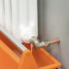 Biały kaloryfer, miedziana rura i pomarańczowy pojemnik na wodę.