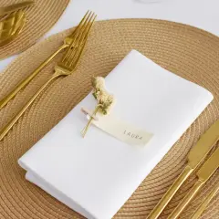 Elegante Tischdeko: Weiß gefaltete Servietten mit Namen "Laura", goldenes Besteck und kleine Trockenblumen auf einem geflochtenen Platzset.
