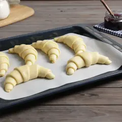 Sześć surowych rogalików z ciasta francuskiego czeka na pieczenie. Obok słoiczek z dżemem.