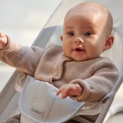 Uśmiechnięte niemowlę w beżowym sweterku siedzi w leżaczku BabyBjörn. Ten leżaczek jest idealny od pierwszych miesięcy życia dziecka.