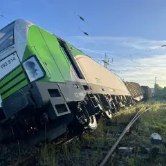 Zielony pociąg towarowy wykoleił się, przechylając się na bok na torach. W tle błękitne niebo i linie energetyczne.