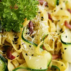 Pyszny makaron z cukinią i boczkiem, posypany parmezanem i czarnym sezamem, udekorowany natką pietruszki.