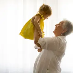 Uśmiechnięta babcia podrzuca w górę wnuczkę w żółtej sukience. Radość i babciowe chwile.