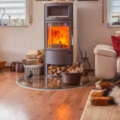Un poêle bouilleur mixte crépite, réchauffant le salon où un chien dort paisiblement sur un tapis. Bois et paniers complètent ce tableau cosy.