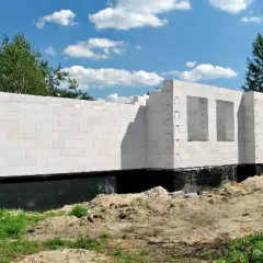 Nowe, białe ściany domu z bloczków betonowych, z otworami na okna i drzwi, stoją na czarnym fundamencie.
