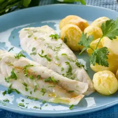 Dwa filety ryby na parze, posypane natką pietruszki, podane z młodymi ziemniakami na niebieskim talerzu.