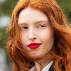 Eine Frau mit leuchtend roten Haaren, die im Wind wehen. Ihr Blick ist nach rechts gerichtet, ihre Lippen sind rot geschminkt. Natur und rote Haare vereinen sich in diesem Porträt.