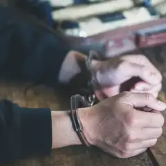 Ręce w kajdankach na drewnianym stole, w tle walizka z paczkami.