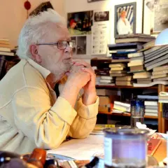 Älterer Mann mit Brille und weißem Haar sitzt am Schreibtisch, umgeben von Büchern.