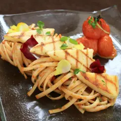 Makaron z halloumi, grillowane plastry sera, pomidorki koktajlowe i listki ziół na szklanym talerzu.
