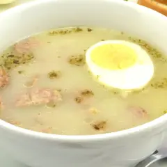 Biała miska z zupą, w której widać kawałki kiełbasy i połówkę jajka na twardo.