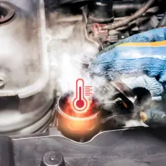 Para mechanika w rękawicy próbuje otworzyć korek chłodnicy, z którego wydobywa się para. Widoczny symbol termometru wskazuje na **wysoką temperaturę płynu chłodniczego**.