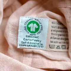 Metka z symbolem GOTS Organic na różowej tkaninie. Informuje o ekologicznym pochodzeniu materiału i zaleca delikatne pranie, bez suszenia w suszarce bębnowej.