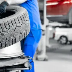 Mechanik w niebieskim kombinezonie trzyma oponę na tle warsztatu samochodowego.