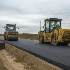 Dwa żółte walce drogowe, każdy to wolnobieżny pojazd budowlany, ubijają świeży asfalt na nowej drodze.