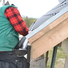 Montaż rynny plastikowej na altanie. Pracownik w kamizelce i flanelowej koszuli montuje czarną rynnę do drewnianej konstrukcji dachu.