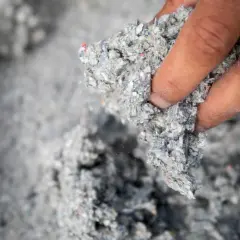 Dłoń trzyma grudkę szarej, włóknistej masy. To materiał izolacyjny z recyklingu, oparty na celulozie.