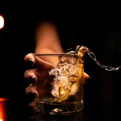 Dłoń trzyma szklankę z whisky, w której widać bryzgający płyn. Ile wodka ma weglowodanow? Zero, to czysty alkohol.