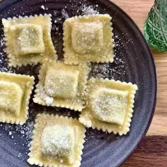 Pięć kwadratowych ravioli z ricottą, posypanych parmezanem, na ciemnym talerzu. Obok zielony kieliszek.