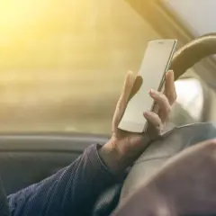 Ręka trzymająca telefon podczas jazdy samochodem. Słońce świeci przez szybę, oślepiając kierowcę.