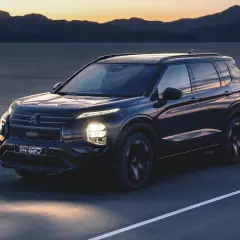 Ciemny Mitsubishi Outlander PHEV jedzie drogą o zachodzie słońca, z górami i jeziorem w tle.