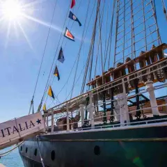La nave Italia, con le sue vele spiegate e le bandiere colorate, si staglia contro il cielo azzurro.