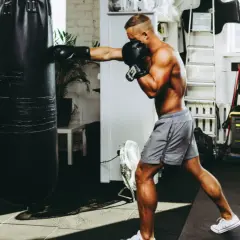 A man boxing with a heavy bag in a gym. He's wearing boxing gloves and shorts, focused on his training.