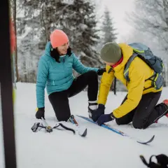 Para przygotowanie do sezonu narciarskiego, mężczyzna w żółtej kurtce pomaga kobiecie założyć narty biegowe w zaśnieżonym lesie.