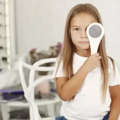 Dziewczynka zasłania oko przyrządem optometrycznym. W tle widać półki z okularami. Może to być badanie wzroku, które czasem wymaga atropiny, a jej skutki uboczne są dobrze znane.