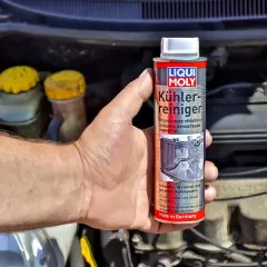 Ręka trzyma puszkę preparatu do płukania układu chłodzenia Liqui Moly. W tle widać silnik samochodu.