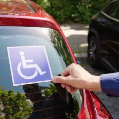 Ręka przykleja niebieską naklejkę z symbolem osoby na wózku inwalidzkim na tylnej szybie czerwonego samochodu. To przypomnienie o tym, jakie dokumenty do karty parkingowej są potrzebne.