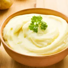 Un tazón de cremoso puré de patatas, decorado con perejil, junto a patatas crudas. ¡Así es como hacer puré de patatas perfecto!