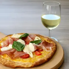 Pizza con prosciutto, mozzarella e basilico su tagliere di legno, accanto a un bicchiere di vino bianco. Un perfetto abbinamento vino pizza.
