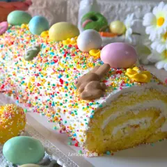 Bûche roulée décorée pour Pâques, recouverte d'un glaçage mascarpone facile et de vermicelles colorés, avec des œufs et un lapin en chocolat.