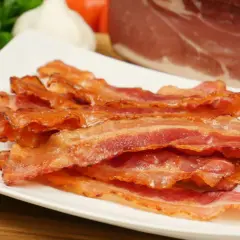 Knusprig gebratener Bacon im Ofen, auf einem weißen Teller gestapelt. Im Hintergrund ein Stück rohes Fleisch, Knoblauch und Petersilie.