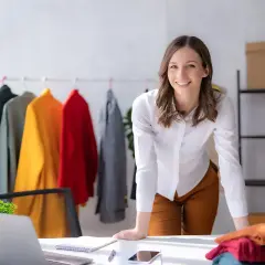 Une chef de produit textile souriante, entourée de vêtements colorés sur un portant, travaille dans son bureau lumineux.